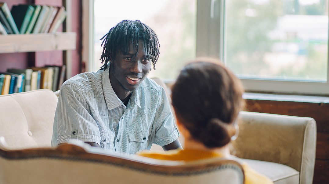 Two individuals are seated in a comfortable room with bookshelves and a large window in the background. The setting suggests a counseling or therapy session, with one person attentively listening to the other. 