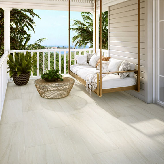 Patio with white stone look rectangle floor tile, swinging couch, wicker table, and view of coast in background.