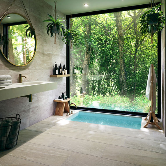 Bathroom with large window wall showing greenery, gray rectangle stone look wall tile, an in floor bathtub, circle mirror, and light gray rectangle stone look floor tile.