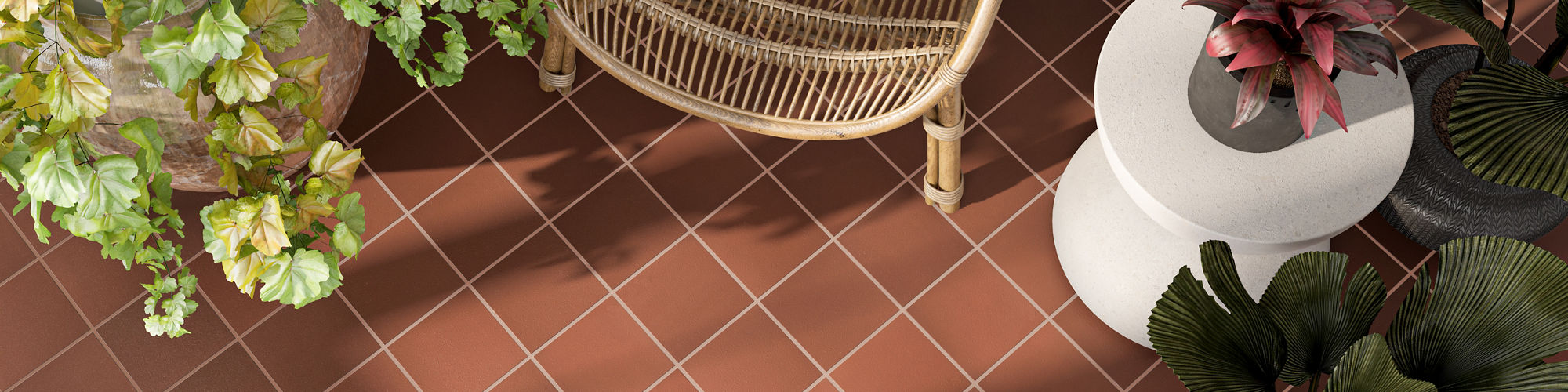 A woven rattan chair sits on a terracotta-colored tiled floor viewed from above. The seating area is surrounded by lush potted plants with green, purple, and pink foliage. Sunlight casts soft shadows across the tiles, enhancing the geometric grid pat
