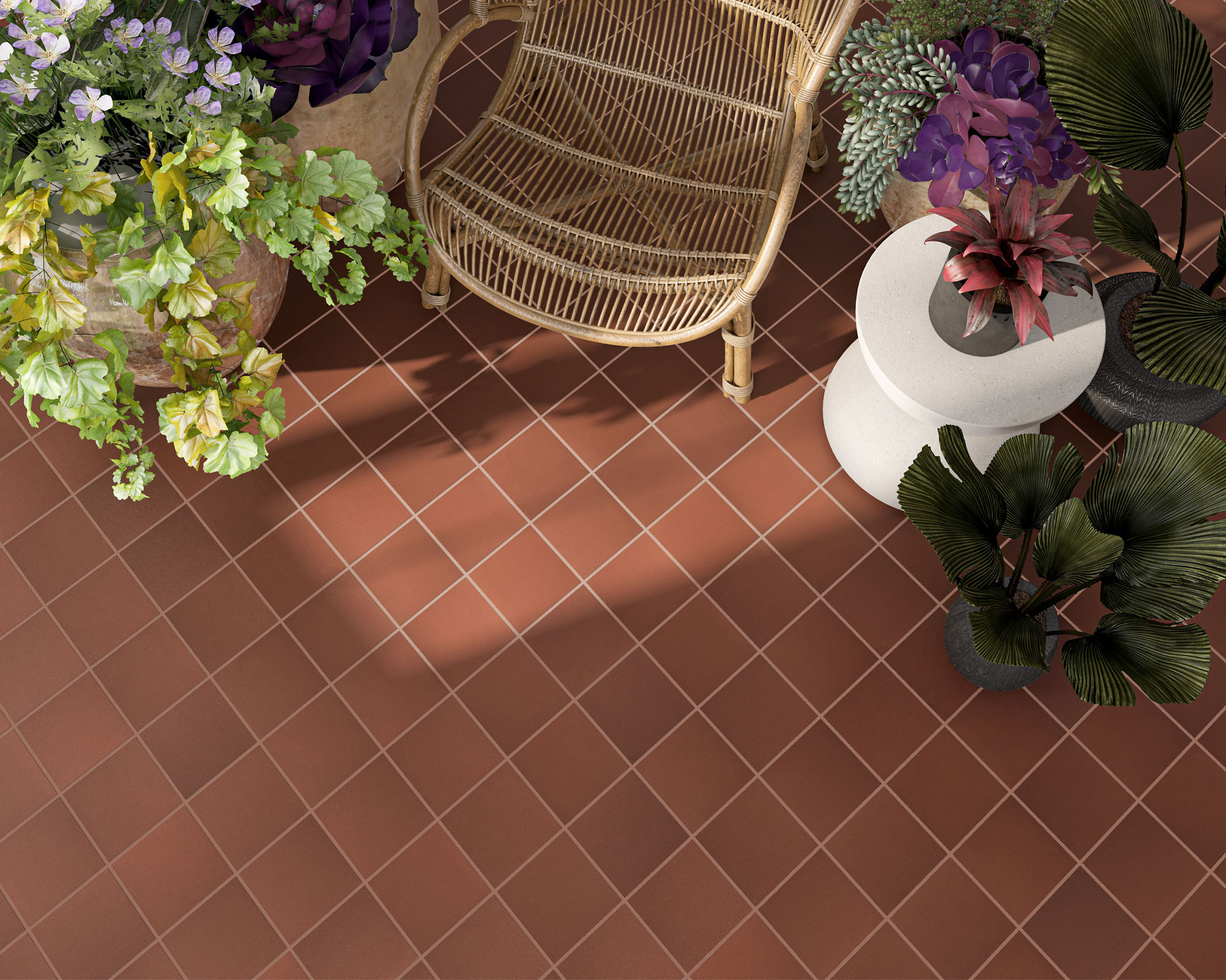 A woven rattan chair sits on a terracotta-colored tiled floor viewed from above. The seating area is surrounded by lush potted plants with green, purple, and pink foliage. Sunlight casts soft shadows across the tiles, enhancing the geometric grid pat