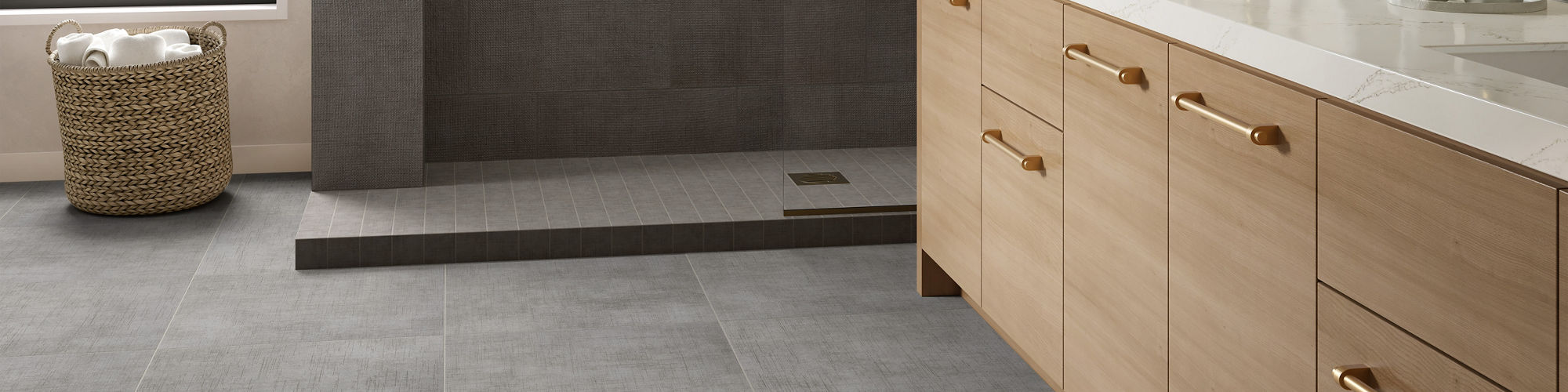 A spacious modern bathroom features large gray floor tiles and a matching gray-tiled walk-in shower. A light wood vanity with white countertop and gold hardware runs along one wall, topped with an arched mirror and wall-mounted fixtures. Natural ligh