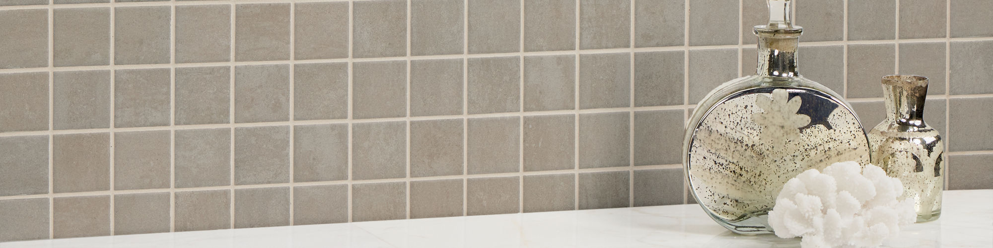 Taupe concrete look square mosaic on wall, and two glass bottles of pebbles on the side.