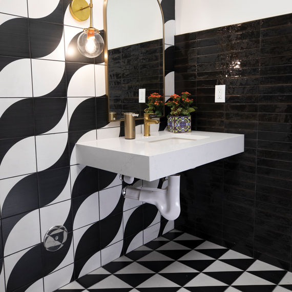 A contemporary bathroom features a floating white sink against a striking tiled wall. Large black and white wave-pattern tiles contrast with glossy black rectangular tiles on the adjacent wall. The floor continues the graphic theme with a black and w