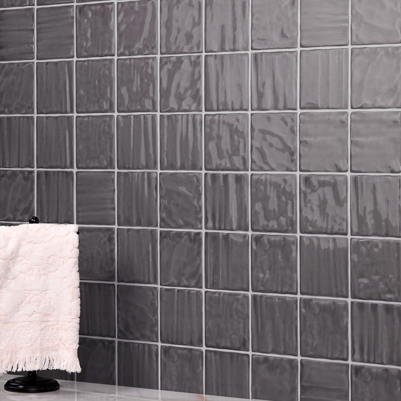A wall covered in glossy gray square tiles forms the main background. The tiles have a slightly wavy, reflective surface that adds texture and depth. In the foreground, a soft light-colored towel hangs on a black stand placed on a smooth countertop. 
