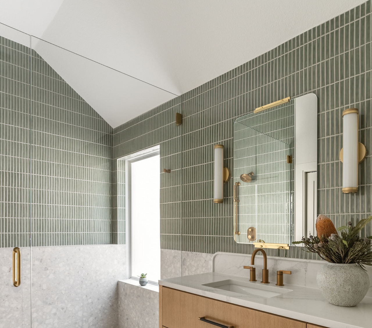 Bathroom with green subway tile on walls and top half of shower wall, light wood cabinets, white marble look countertop, and white terrazzo look tile on bottom half of walls and shower wall.