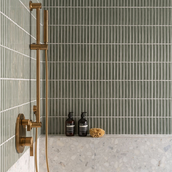 Close up of shower with green rectangle mosaic on shower walls, white terrazzo look tile on bottom half of wall, and gold accents.
