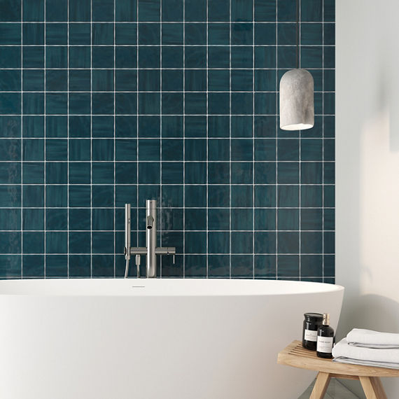 A contemporary white freestanding bathtub is set against a wall of glossy teal square tiles. A sleek metal faucet rises from the tub edge, emphasizing minimalist bathroom design. A small wooden stool holds dark bottles and a candle, adding spa-like d