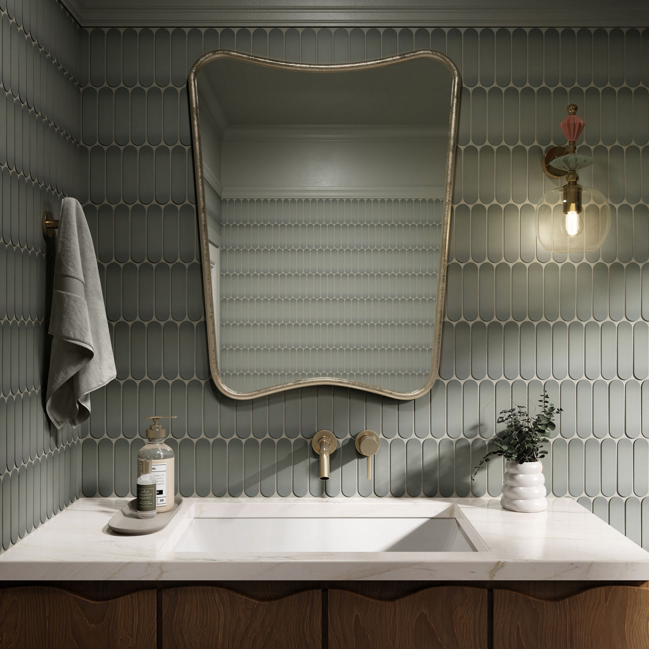 A contemporary bathroom features a unique curved mirror above a white marble countertop and wooden vanity. The wall is covered in elongated oval tiles in a muted green shade, complemented by a brass wall sconce with an exposed bulb. A towel hangs to 