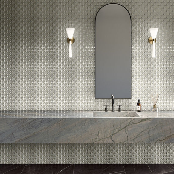 A contemporary bathroom features a long stone vanity with an integrated sink and sleek faucet. The wall behind the vanity is covered in light-colored, geometric textured tiles that create a subtle three-dimensional pattern. An arched mirror is center
