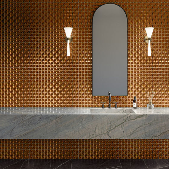 A contemporary bathroom interior features a long stone countertop and integrated sink against a striking copper-toned textured tile wall. An arched mirror is centered above the basin, flanked by two sleek wall sconces providing warm ambient light. Th