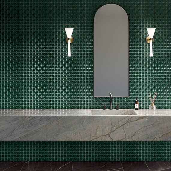 Contemporary bathroom interior featuring a long stone countertop and integrated sink. A tall arched mirror is centered on a dark green textured tile wall. Two wall sconces with white shades and brass mounts flank the mirror, providing soft lighting. 