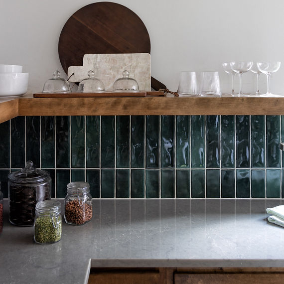 Kitchen with blue rectangle tile on backsplash, gray stone look porcelain countertop slab, wood cabinets, wood shelf, and dishes on shelf.