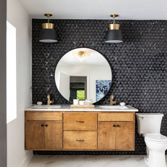 Bathroom with a black triangle mosaic on the walls, wood cabinets, white countertop, and a round mirror.