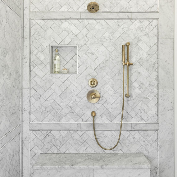Shower wall with white and gray veined marble tile in a herringbone pattern, shower niche, shower bench, and matte brass shower fixtures.