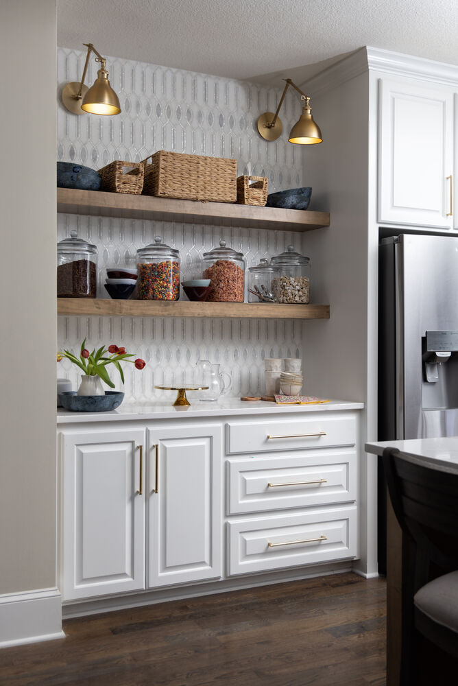 A contemporary kitchen pantry nook features white cabinetry with gold hardware beneath two floating wooden shelves. Clear glass jars filled with dry goods and snacks are neatly arranged on the shelves alongside woven baskets. Brass wall sconces provi