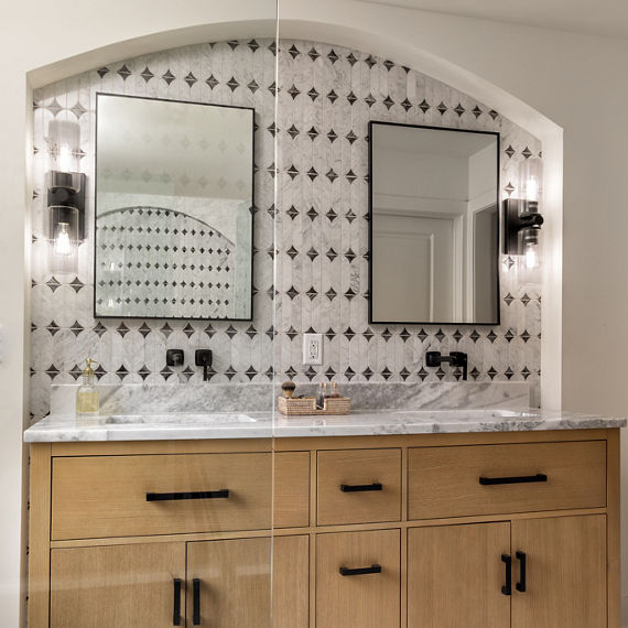 Bathroom with light brown wood cabinets on his and hers sinks, white marble floor, white marble look mosaic on wall, and white marble slab on the countertops.