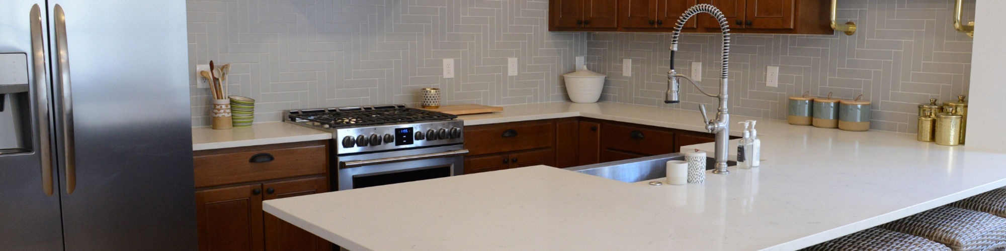 Remodeled kitchen with gray wall tile in a herringbone pattern, white quartz countertop and peninsula, natural wood cabinets and vent hood, and floating shelves.