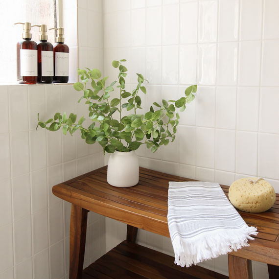 Shower with wooden bench, white vase with greenery, white subway tiles on walls, and gold accents.