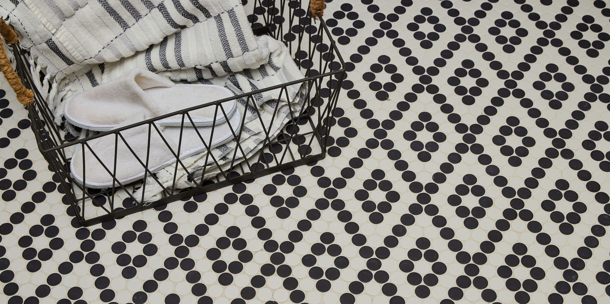 Close up of bathroom with black and white penny round mosaic in diamond pattern on floor, black basket, and towels inside basket.