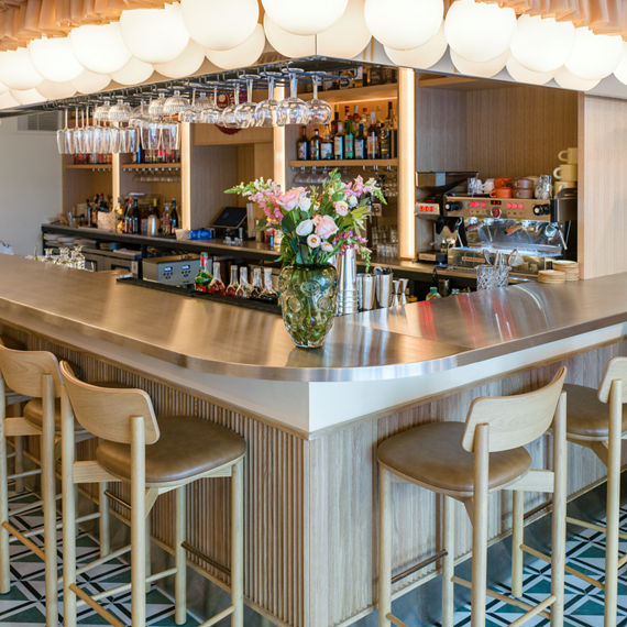 A contemporary bar features a sleek wooden counter with light wood stools arranged around it. The setting is highlighted by a striking ceiling installation of draped fabric and globe pendant lights, creating a warm and inviting atmosphere. Glassware 