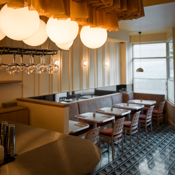 A stylish cafe interior features round globe pendant lights and a unique ceiling treatment above a bar area. The seating area includes cushioned booths and wooden chairs arranged around white tables, set against a backdrop of vertical wall panels. Th