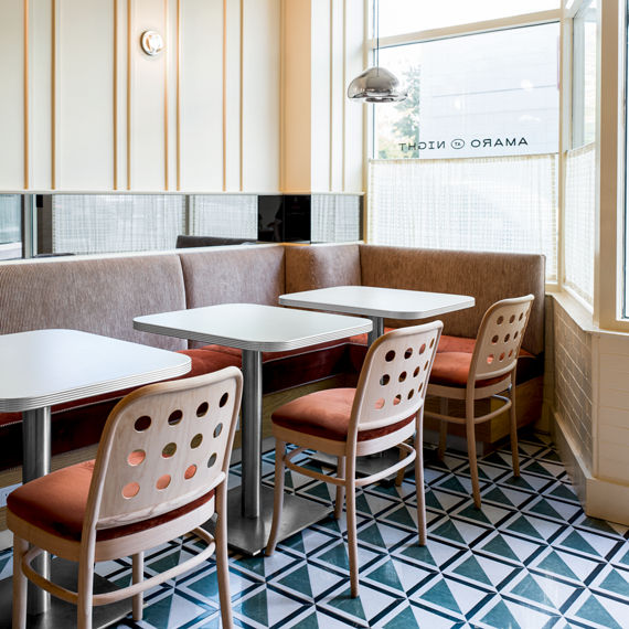 A cozy corner booth in a modern cafe features light wood paneling, geometric blue and white floor tiles, and minimalist tables. Four chairs with circular cutouts and orange cushions surround the tables, while large windows allow natural light to fill