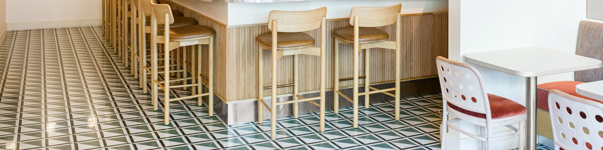 A contemporary cafe features a central bar area with wooden stools and a geometric patterned tile floor. The bar is accented by a row of globe pendant lights and a draped fabric canopy, creating a cozy and inviting atmosphere. The setting includes fr