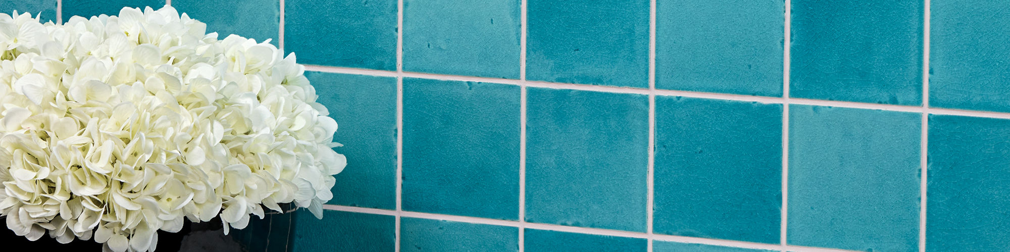 Light blue square tile on wall with handcrafted look, and black pot with white flowers on the side.