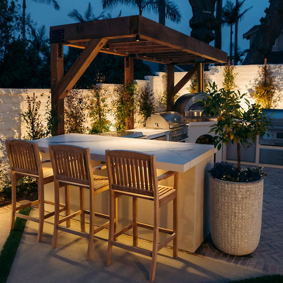 Outdoor kitchen with white marble look porcelain counter, wooden chairs, and a silver grill.