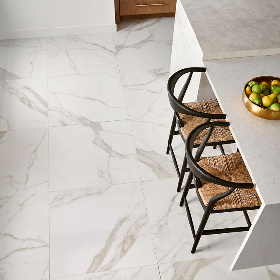 Over head view of kitchen with white marble look quartz square floor tile, white island, wicker chairs, and golden bowl of fruit.