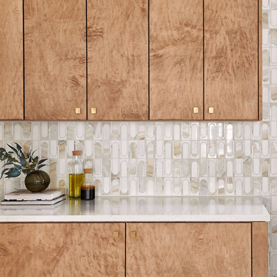 Kitchen with wood cabinets and accents, two islands, white marble look waterfall countertops, white mosaic on backsplash, and black island chairs.