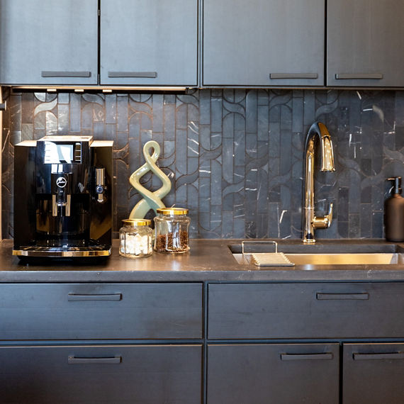 Kitchen with black natural stone mosaic on backsplash, black marble look countertop, black cabinets, and coffee machine.