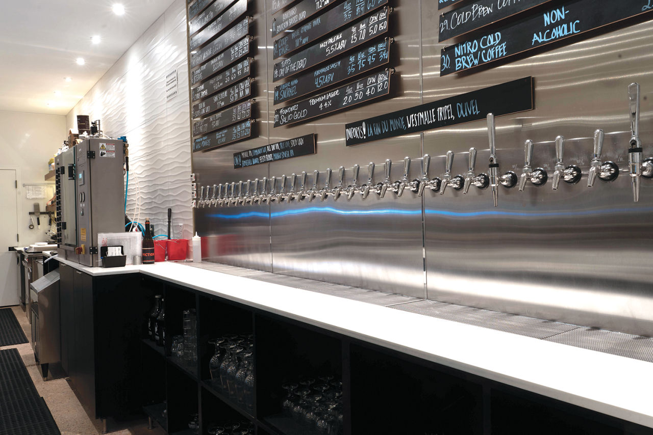 A sleek taproom interior features a long row of stainless steel beer taps set into a reflective metal wall.