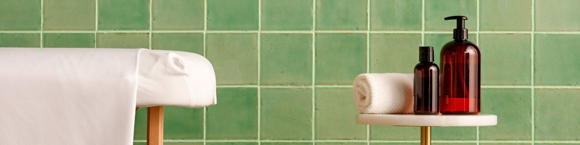 Spa with light green square tile on wall, small table with lotion and towel, and massage table.