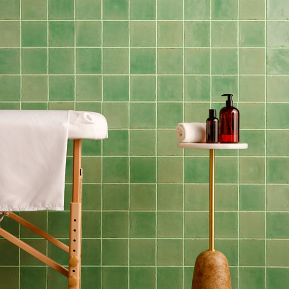 Spa with light green square tile on wall, small table with lotion and towel, and massage table.