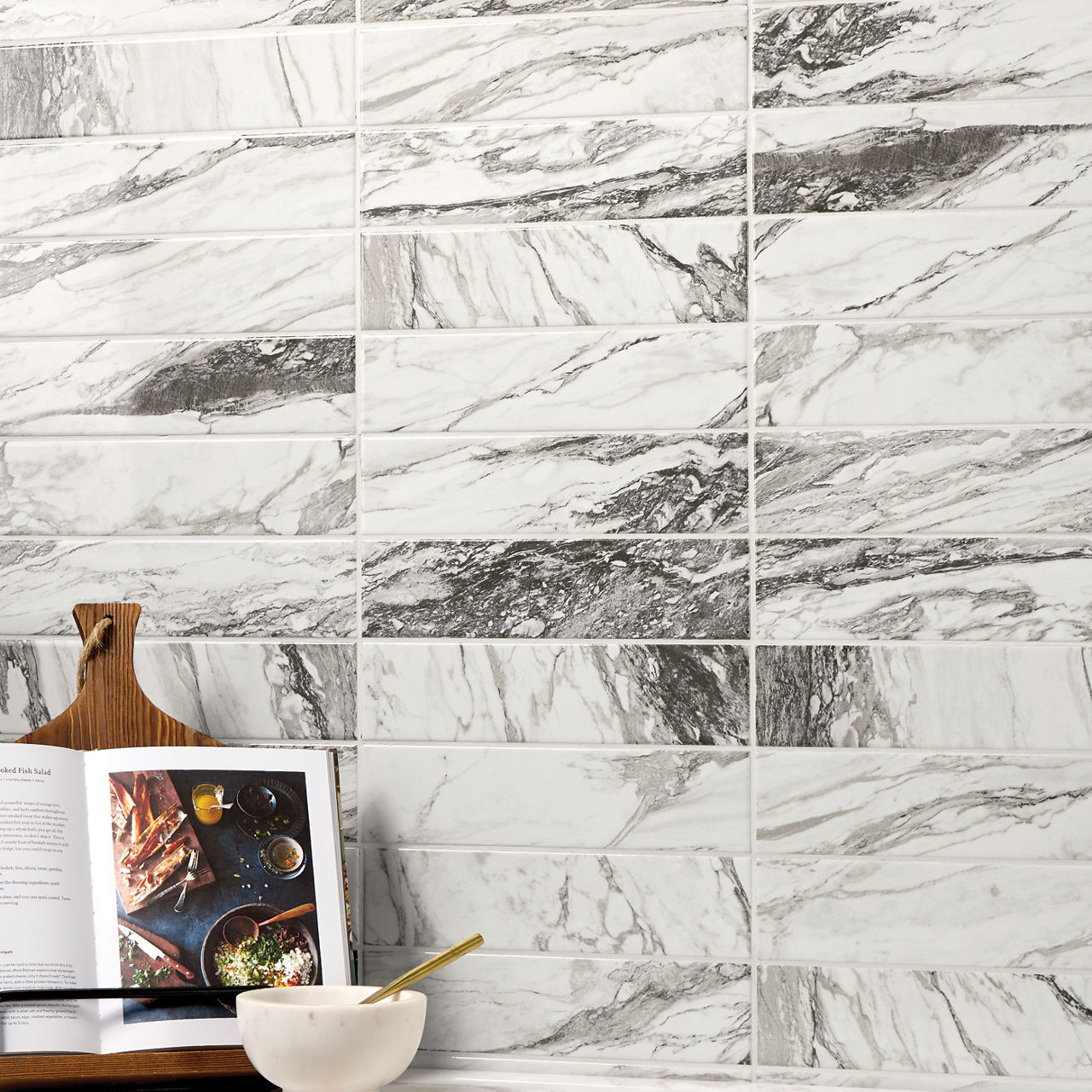 The image features a wall covered in rectangular marble-look tiles with gray and white veining. In the foreground, there is an open cookbook displayed on a wooden board, accompanied by a white bowl and a golden spoon. The setting suggests a modern ki