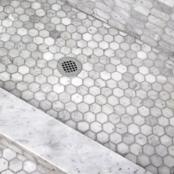 Close up of shower floor with white marble hexagon mosaic on floor and white marble elongated hexagon mosaic on wall.