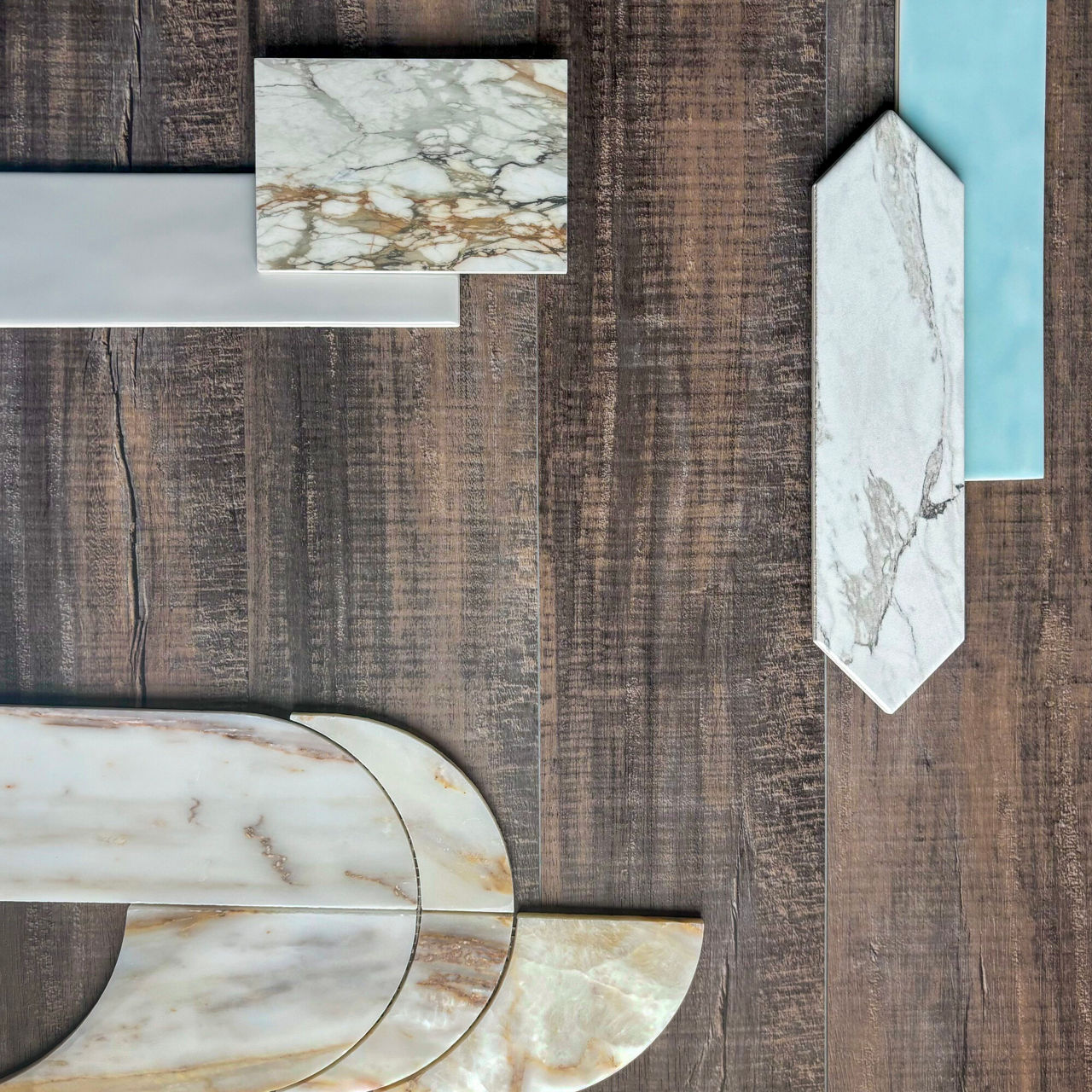 A collection of decorative tiles is arranged on a rustic wooden surface. The tiles include rectangular, curved, and elongated hexagonal shapes in marble and smooth ceramic finishes. Colors range from white and cream with brown veining to light blue a