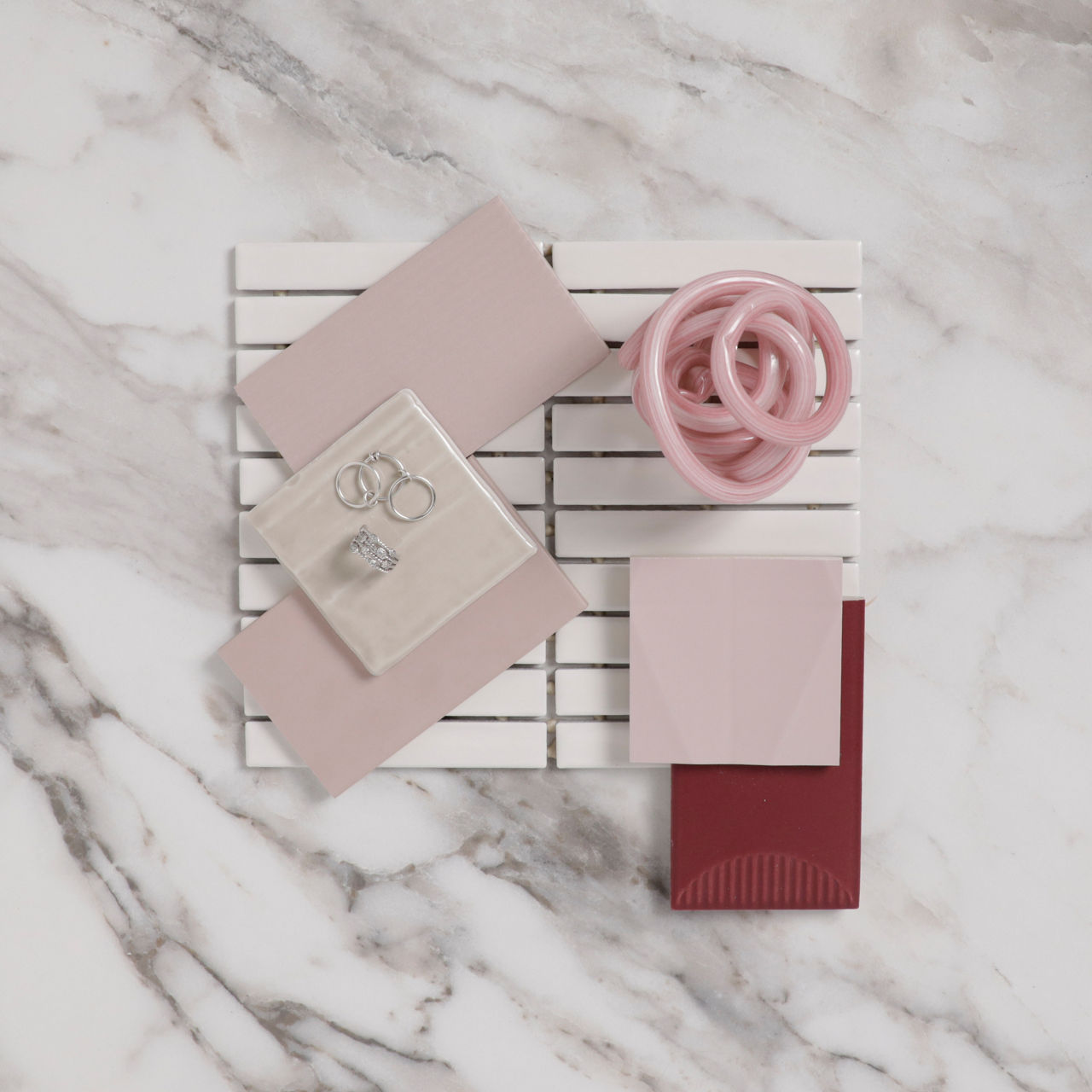 Flatlay with light pink rectangle tile, a large white marble look tile, a dark pink tile with a half moon design, small pink square tile, and silver jewelry on flatlay.