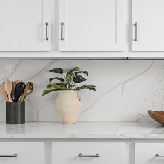 Close up of kitchen with white marble look countertops and backsplash, white cabinets, wooden bowl on counter, and vase with greenery.
