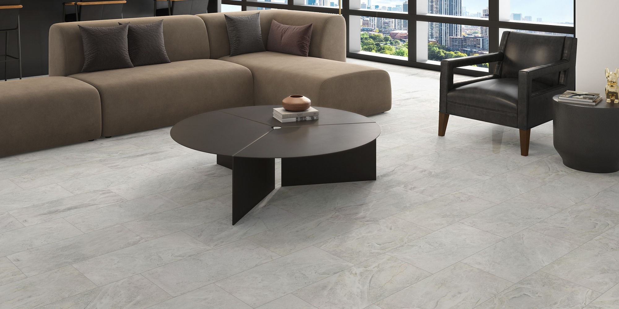 Contemporary living room features a large sectional sofa and a round coffee table on light stone-look flooring. Floor-to-ceiling windows reveal a bright city skyline, filling the space with natural light. A sleek black armchair and minimalist decor c