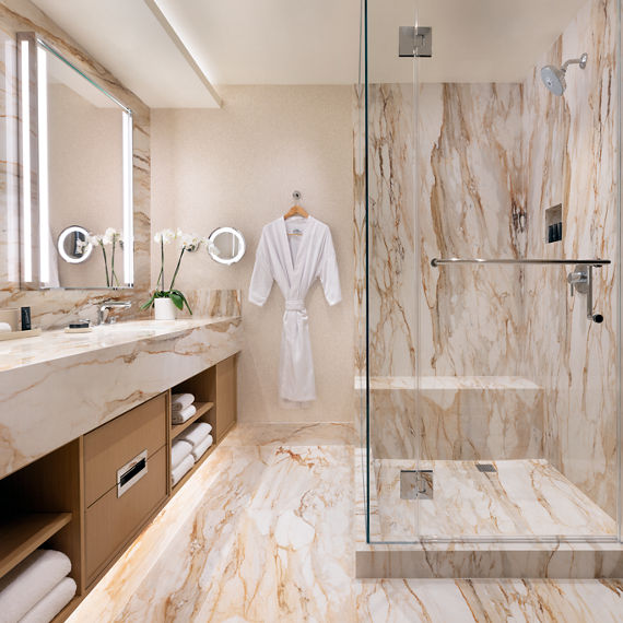 A spacious, luxurious bathroom features extensive marble surfaces on the floor, walls, and countertop. A large glass-enclosed walk-in shower stands to the right with a metal grab bar and rainfall showerhead. On the left, a long vanity with dual sinks