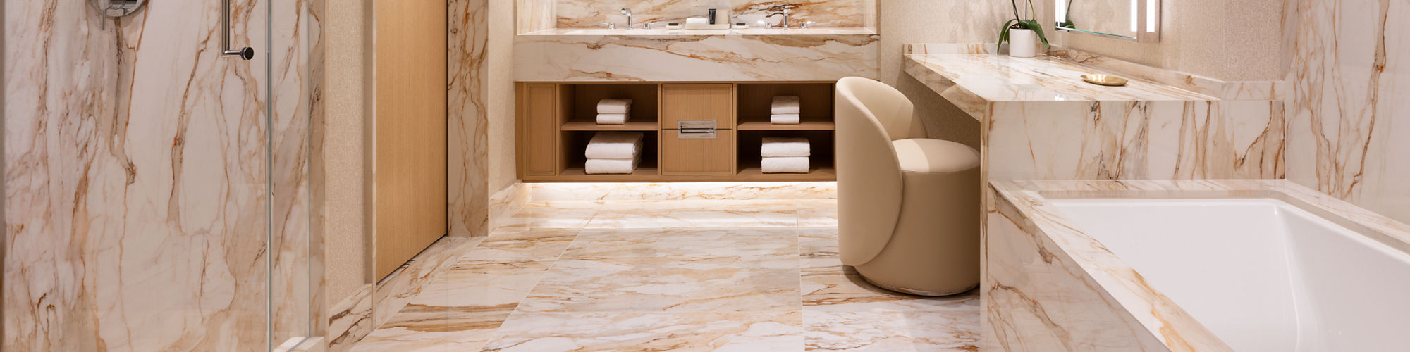 Spacious contemporary bathroom featuring extensive beige and white marble surfaces. A large vanity with double mirrors and integrated lighting sits opposite a built-in bathtub. A glass-enclosed shower and wooden door are visible on the left side. Sof