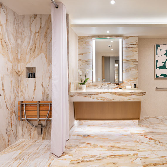 A spacious bathroom features extensive marble walls and flooring with warm beige and gold veining. On the left, a walk-in shower includes a handheld showerhead, grab bars, and a fold-down wooden bench behind a light curtain, highlighting accessibilit