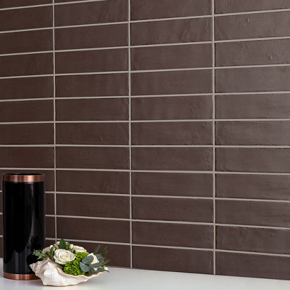 A sleek brown rectangular tile wall forms the backdrop of a minimalist interior scene. In the foreground, a glossy black cylindrical container with metallic copper accents stands on a white surface. Beside it, a decorative shell-shaped dish holds whi
