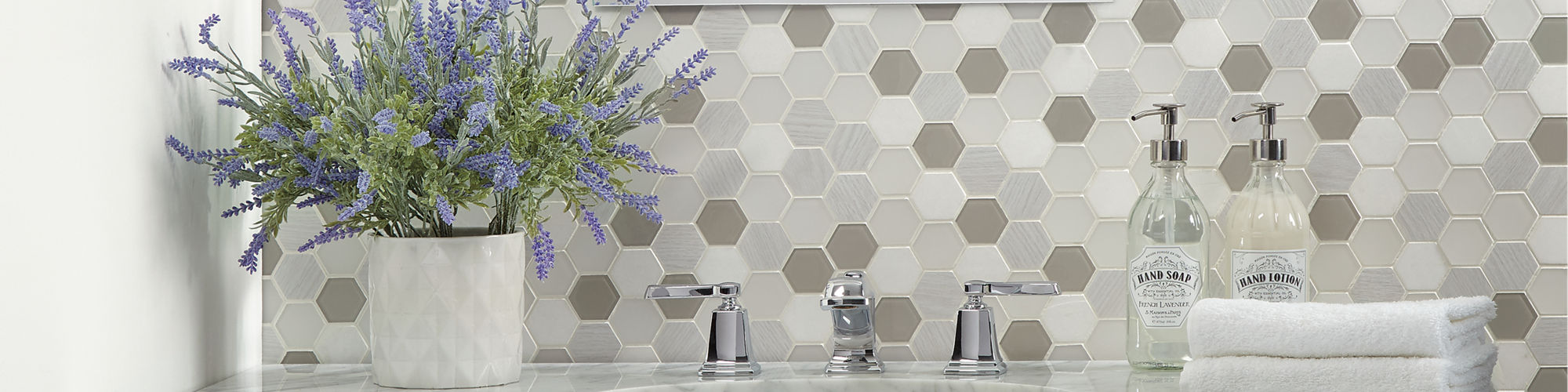 Bathroom with white and gray hexagon mosaic on backsplash, white marble look counter, dark wood cabinets, and white pot with greenery on sink.