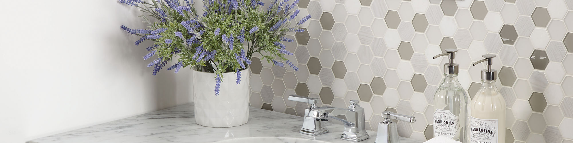 Bathroom with white and gray hexagon mosaic on backsplash, white marble look counter, dark wood cabinets, and white pot with greenery on sink.