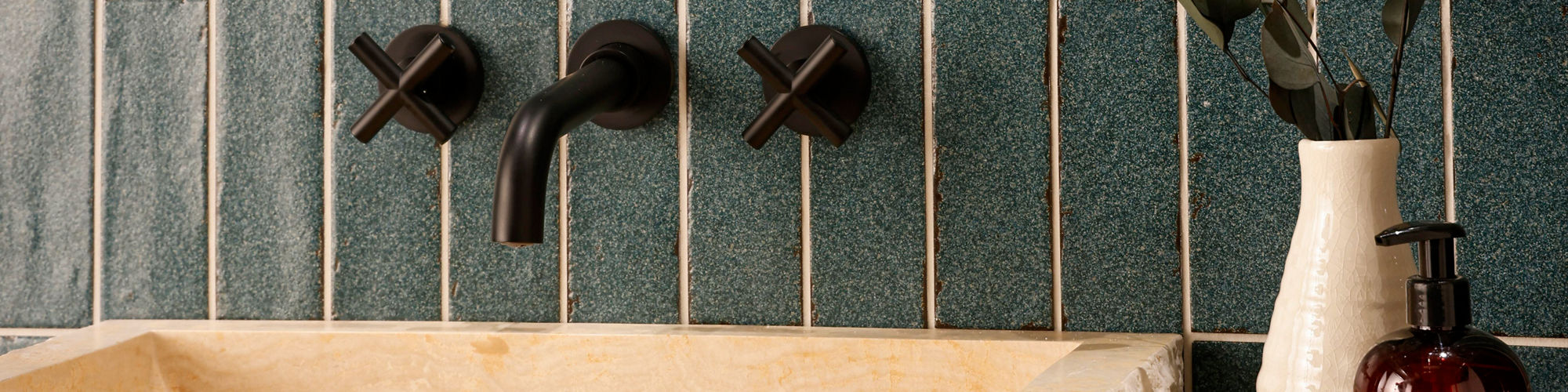 A rectangular stone sink sits atop a marble countertop, set against a wall of vertically arranged blue tiles. Matte black wall-mounted faucet handles add a contemporary touch. A ceramic vase with eucalyptus branches and a brown pump bottle labeled 'A