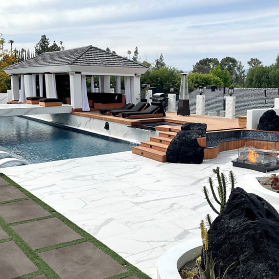 Jason Derulos backyard with pool deck and bridge made of white and gray porcelain tile that looks like marble, and wood deck with lounge chairs.